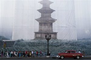 Personas en una parada de transporte con una fotografía del artista visual sudcoreano Han Sungpil, sobre la fachada de un edificio en el centro de La Habana, Cuba, el viernes 22 de mayo de 2015. La fotografía es parte de la Bienal de Arte de La Habana. (Foto AP/Desmond Boylan)